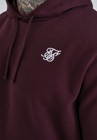 Felpa con cappuccio color marrone scuro, dal texture morbido, con un logo bianco ricamato sul petto e senza hardware o dettagli visibili.
