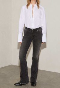 Chemise blanche à col froncé et manches longues, associée à un jean noir évasé avec des ourlets effilochés et un délavage clair. Bottines noires.