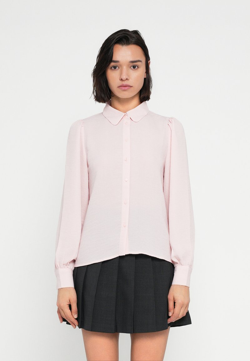 Ροζ ανοιχτό πουκάμισο με γιακά, μακριά φουσκωτά μανίκια και ελαφρώς κυρτή ρέλι, συνδυασμένο με μια πλισέ σκούρα φούστα.