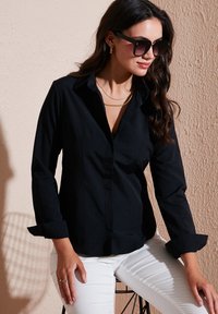 Black button-up shirt with a collar, long sleeves rolled at the cuffs, paired with white pants; model wearing large black sunglasses and a gold necklace.