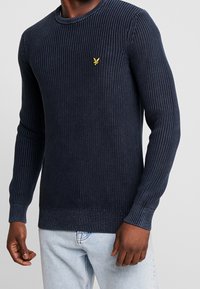 Marineblauer, gerippter Strickpullover mit langen Ärmeln und Rundhalsausschnitt. Mit einem kleinen gelben Logo auf der Brust. Bequeme, figurbetonte Passform.