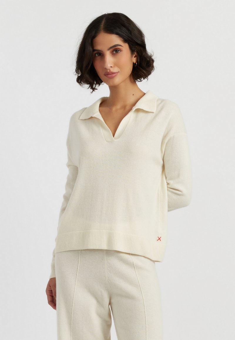 Cremefarbener Strickpullover mit V-Ausschnitt und Kragen, mit geripptem Saum und Bündchen. Rote Akzentdetails an der Seite. Weicher, strukturierter Stoff.