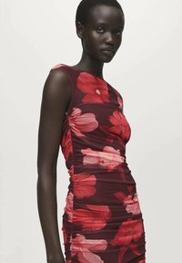 Robe moulante d'un bordeaux foncé avec de grands motifs floraux rouges et roses. Comprend des côtés froncés et un design sans manches.