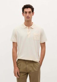 Polo de color beige claro hecho de algodón suave, con un cuello corto, tres botones y un bolsillo en el pecho. Combinado con pantalones caqui.