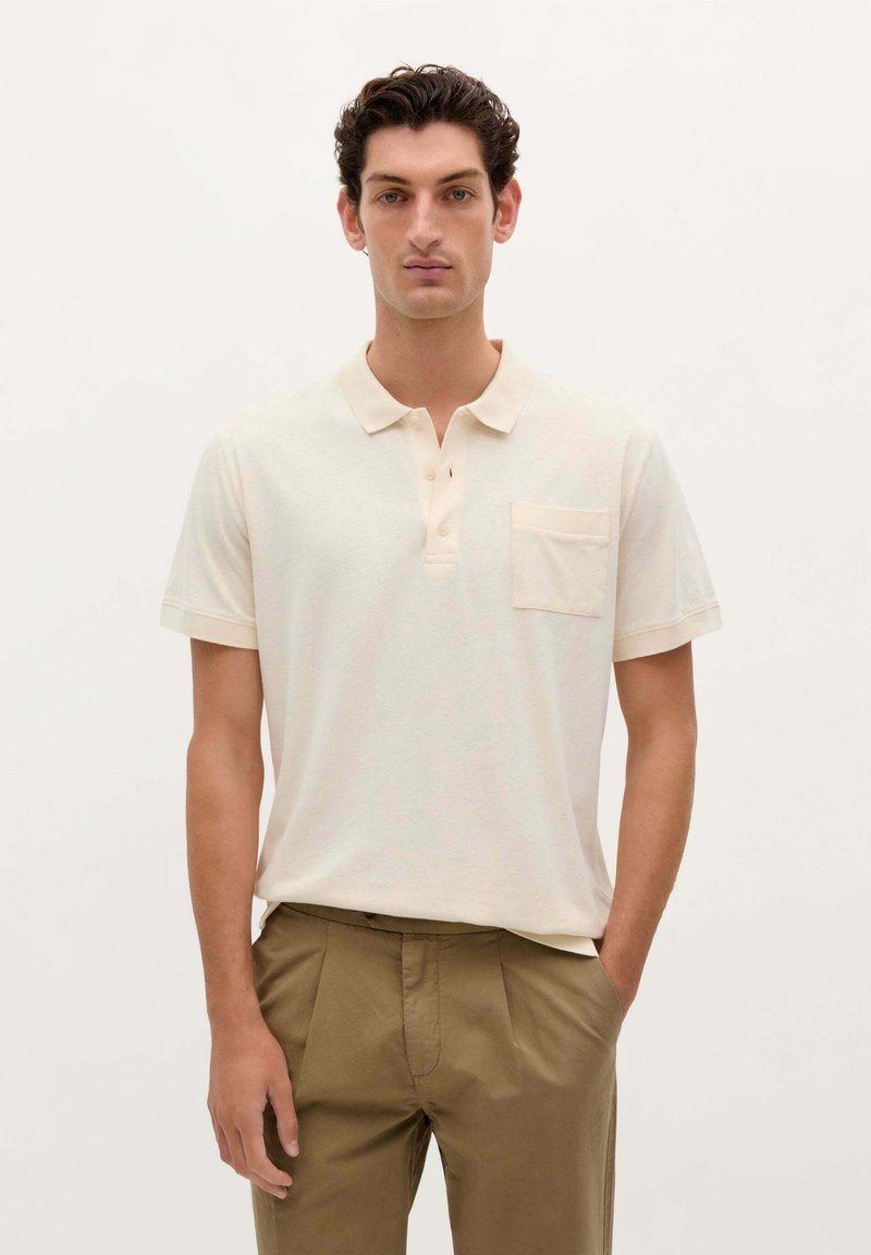 Polo de color beige claro hecho de algodón suave, con un cuello corto, tres botones y un bolsillo en el pecho. Combinado con pantalones caqui.
