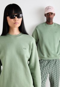 Two models wear coordinating light green sweatshirts; one has a black cat-eye sunglasses, while the other wears patterned pants and a beige cap.