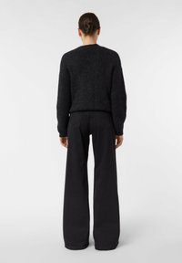 Kobieta z włosami upiętymi w kok, ubrana w czarny prążkowany sweter i szerokie czarne spodnie, stojąca na tle jednolitego białego tła.