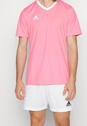 Uomo che indossa una maglietta sportiva Adidas rosa a maniche corte e pantaloncini bianchi Adidas, in piedi davanti a uno sfondo semplice.