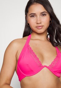 Top de biquíni cor-de-rosa com alças halter, apresentando uma ribana recortada ao longo da gola e um tecido liso e elástico. Design simples e ajustado.