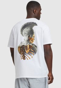 Valge puuvillane t-särk, mille seljal on detailne graafika kiivrit kandvast figuurist, millel on kuldsed skeletielemendid. Lühikesed varrukad, mugav lõige.