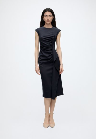 Femme debout dans une robe midi noire ajustée sans manches avec des détails froncés et drapés, portant des talons beiges à bride à la cheville sur un fond uni.