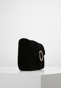 Svart vadderad sammet handväska med detalj i guld cirkulär ring. Har en klaffstängning och slät textur, ligger på en vit yta.