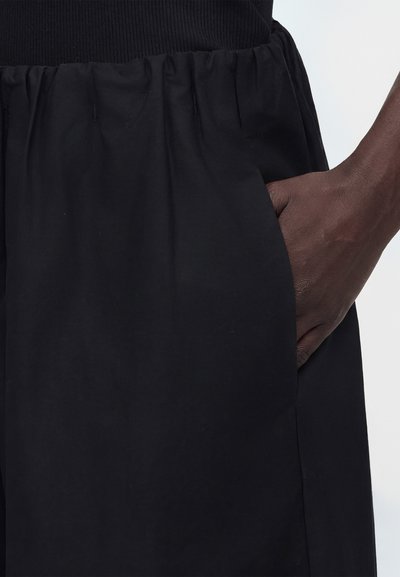 Σκούρο δέρμα χέρι τοποθετημένο μέσα στην τσέπη μιας μαύρης φούστας με ελαστική ζώνη στη μέση, φορεμένη με μαύρο τοπ.