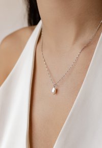 Silberne Kettenhalskette mit gedrehtem Design und einem kleinen weißen Perlenanhänger, gelegen auf einem hellen Stoffhintergrund.