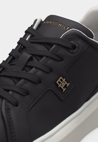 Gros plan d'une basket en cuir noir avec des lacets noirs, un logo Tommy Hilfiger doré sur le côté et des œillets marqués, semelle blanche visible en bas.