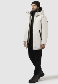 Lys grå parka med hætte, sorte detaljer, front lynlås og trykknaplukninger. Båret over en sort skjorte og sorte cargos, komplementeret af sorte støvler.