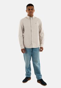 Camicia button-up beige chiaro con colletto, abbinata a jeans azzurri e sneakers nere con dettagli marroni. Il modello è in piedi rivolto verso l'anteriore.