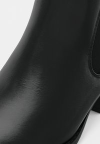 Gros plan d'une botte cheville en cuir noir lisse avec un talon bloc et un panneau latéral élastique.