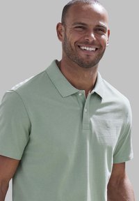 Polo shirt verde chiaro realizzato in tessuto testurizzato, con colletto, due bottoni e un piccolo logo sul lato sinistro del petto. Finitura liscia.