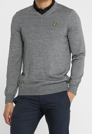 Man draagt een grijze V-hals trui met een klein geel vogellogo op de borst, een zwart shirt eronder en donkerblauwe broek.