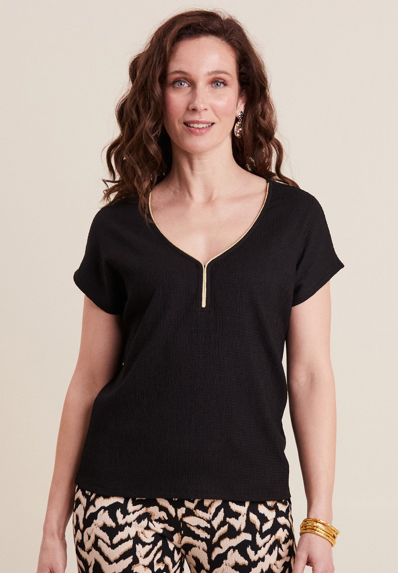 Top nero a maniche corte con scollo a V e dettaglio della cerniera dorata, realizzato in un tessuto strutturato. Abbinato a pantaloni neri e crema con motivo.