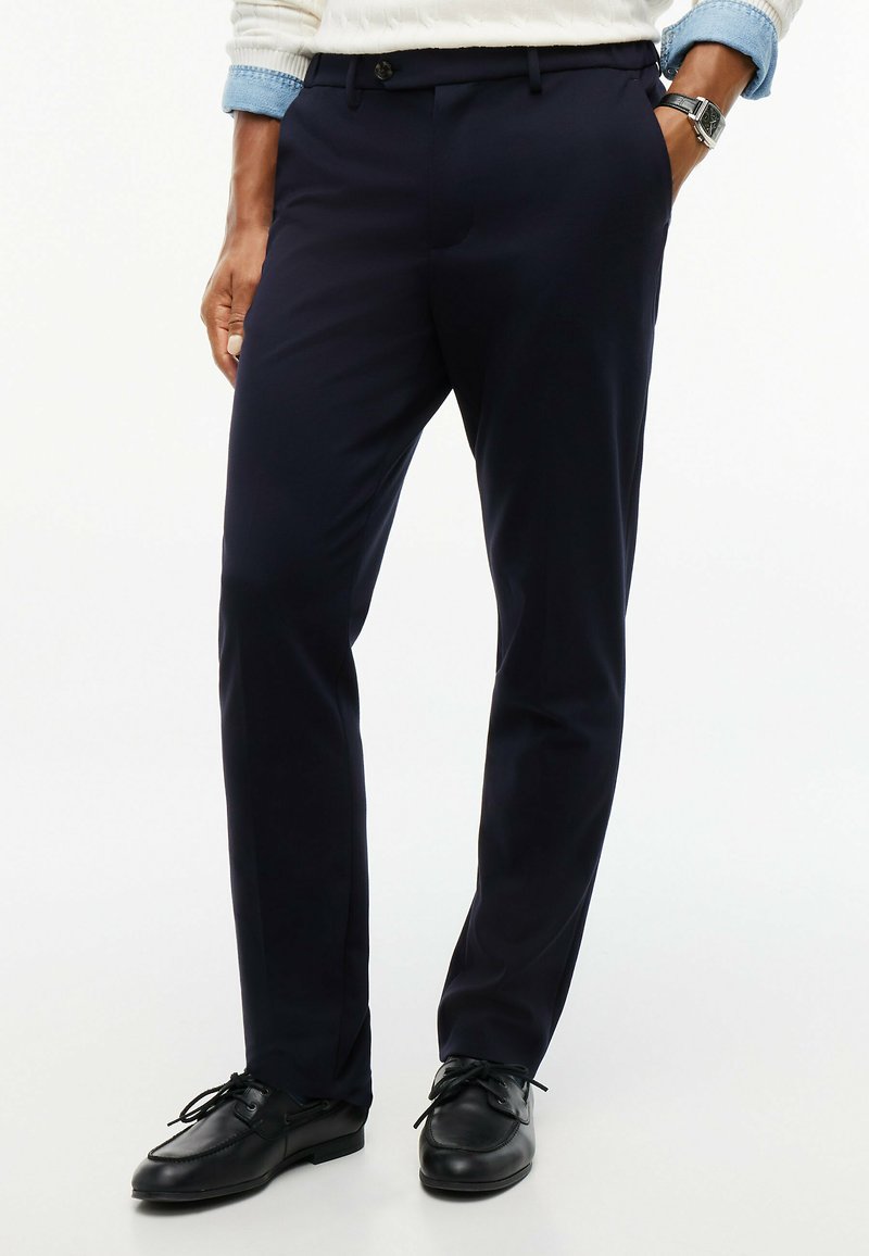 Homme portant un pantalon de costume bleu marine foncé, des chaussures en cuir noires, un pull blanc avec des poignets bleus et une montre-bracelet, une main dans la poche.
