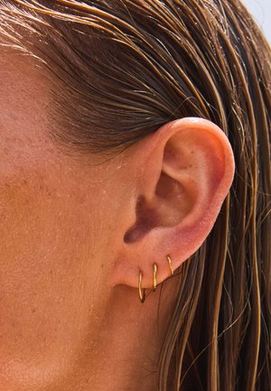 Nahaufnahme eines Ohrs einer Person mit drei kleinen goldenen Creolen und nassem braunem Haar, das hinter dem Ohr liegt.