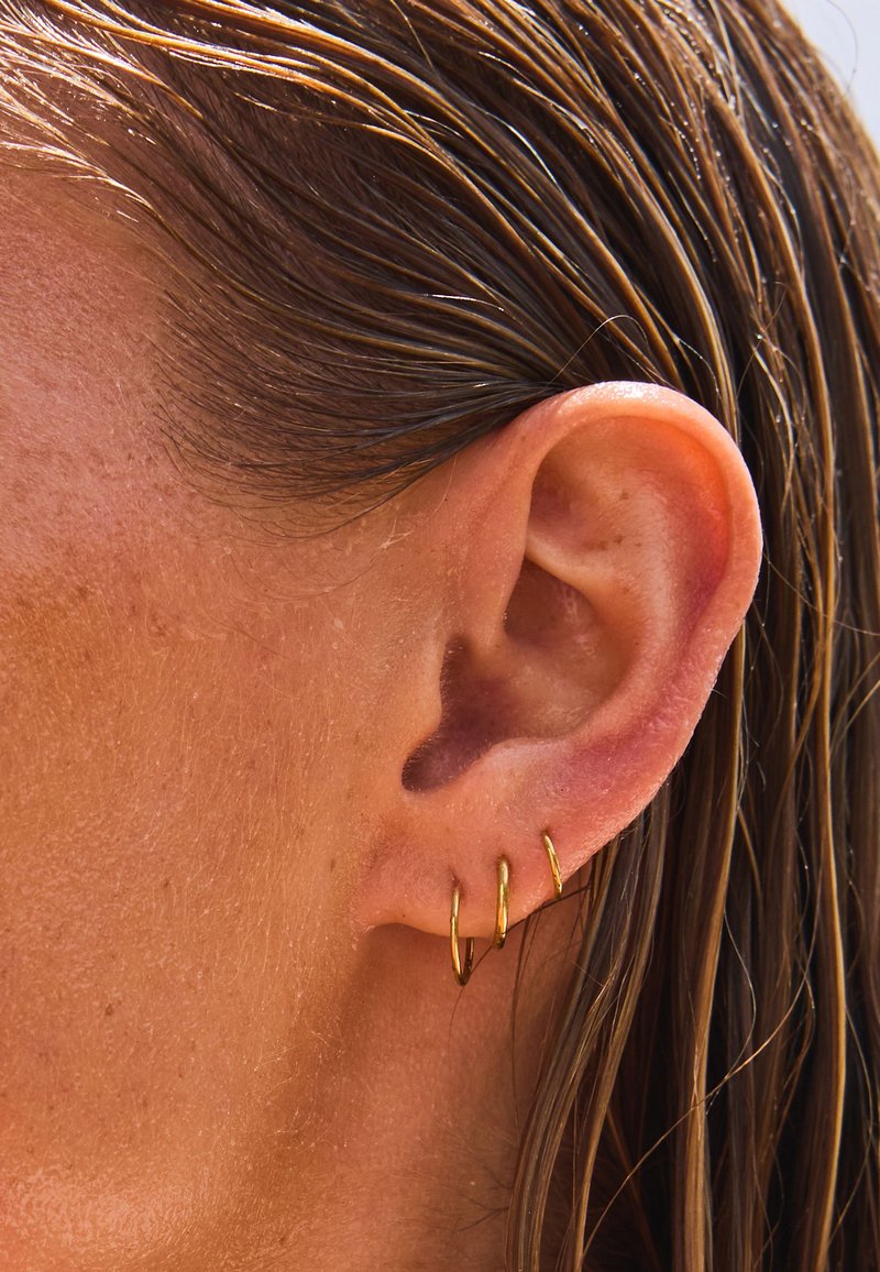 Nahaufnahme eines Ohrs einer Person mit drei kleinen goldenen Creolen und nassem braunem Haar, das hinter dem Ohr liegt.