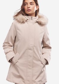 Cappotto invernale beige con cappuccio foderato in pelliccia, chiusura frontale con cerniera e bottoni a pressione. Presenta tasche laterali e un esterno liscio e impermeabile.