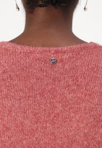 Maglione lavorato a maglia rosa chiaro, con una texture morbida e un ampio scollo. Tag del marchio circolare ben visibile cucito sul retro.
