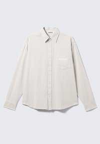 Chemise beige clair à manches longues avec boutons, col classique, poche avant et marquage discret sur la poche en blanc. Tissu à texture lisse.
