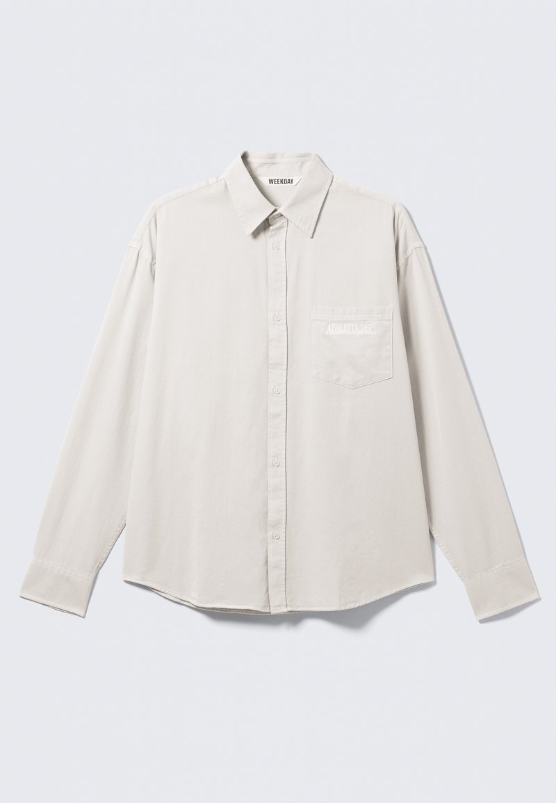 Chemise beige clair à manches longues avec boutons, col classique, poche avant et marquage discret sur la poche en blanc. Tissu à texture lisse.