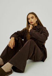 Hnědá denímová košile a široké kalhoty s límcem a zapínáním na knoflíky; modelka sedí, nosí náušnice, texturovaná látka.