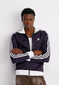 Mornarsko plava adidas zip jakna s bijelim crtama i ovratnikom. Ima srebrni logo na prsima, rebraste manžete i uski kroj.