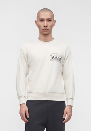 Homme à tête rasée portant un sweat-shirt blanc à manches longues avec le logo « Aries » sur la poitrine et un pantalon noir, debout devant un fond uni.