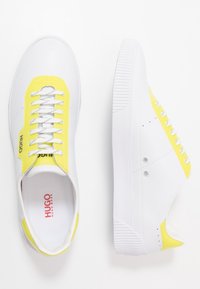 Sneakers bianche con tomaia in pelle liscia e dettagli gialli, lacci piatti e suola in gomma strutturata, progettati con un branding minimale.