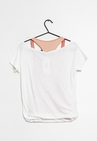 Bílý top s krátkým rukávem a volným střihem, pod ním je vidět broskvově zbarvený tílko, ramínka jsou viditelná a materiál je hladký.