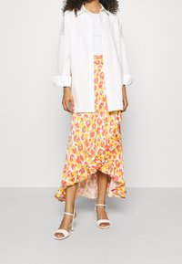 Camicia bianca con bottoni sopra una canotta bianca, abbinata a una gonna maxi con stampa leopardata rosa e gialla caratterizzata da volant e orlo asimmetrico. Sandali bianchi.
