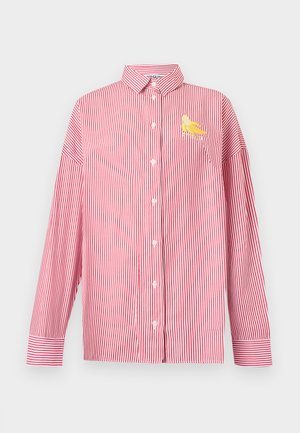 Personne portant une chemise rouge et blanche à rayures oversize par-dessus un tee-shirt blanc, un short en jean au genou avec écritures, des baskets blanches, des chaussettes et des lunettes de soleil noires.