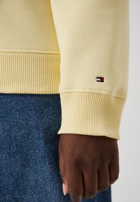 Manche de sweat-shirt jaune avec poignet côtelé ; petit logo avec des rayures rouges, blanches et bleues près de l'ourlet ; associé à un jean bleu.