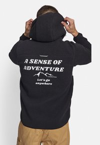 Sudadera con capucha de forro polar negro, con cremallera frontal y cordón en la capucha. La parte trasera presenta texto en blanco: "UN SENTIDO DE AVENTURA" y gráficos de montañas.