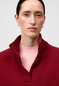 Close-up van een persoon met bruine ogen, die witte creolen draagt, een rode gebreide trui met een hoge kraag en een roze ronde hals T-shirt.