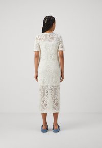 Robe midi en dentelle ivoire avec des manches courtes, des motifs floraux, des bords festonnés et des fentes sur les côtés. Portée avec des sandales à glissière bleues.