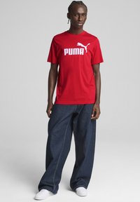 Rotes Baumwoll-T-Shirt mit weißem "PUMA"-Logo und Logo-Grafik. Kombiniert mit lockeren, dunkelblauen Jeans und weißen Turnschuhen.