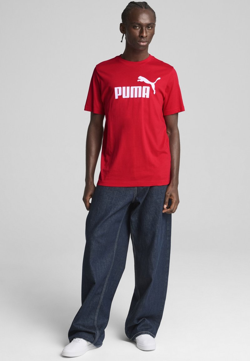Rotes Baumwoll-T-Shirt mit weißem "PUMA"-Logo und Logo-Grafik. Kombiniert mit lockeren, dunkelblauen Jeans und weißen Turnschuhen.