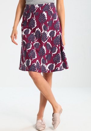 Femme portant une jupe violette mi-longue avec de grands motifs de feuilles blanches et noires, associée à des chaussures beige à bout pointu et sans lacets.
