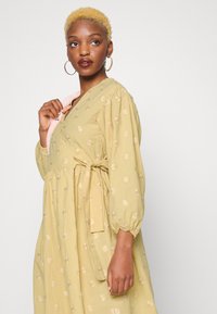 Vestido amarelo bordado com flores, com uma cintura ajustada, mangas bufantes e um detalhe de laço. Tecido de algodão suave com uma textura leve.
