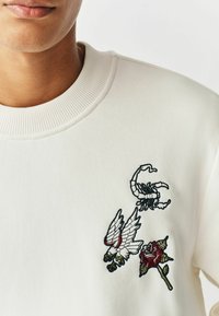 White sweatshirt featuring embroidered designs: a scorpion, wings, and a rose. The fabric has a smooth texture and a ribbed collar.