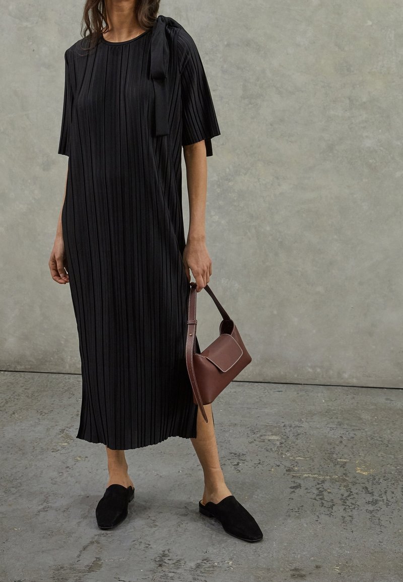 Schwarzes plissiertes Midikleid mit kurzen Ärmeln und einer seitlichen Schleifendetail, kombiniert mit schwarzen Mules und einer braunen strukturierten Handtasche.