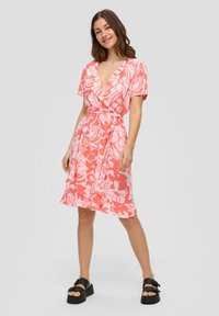 Blumenkleid in hellem Koralle mit kurzen Ärmeln, V-Ausschnitt, tailliertem Schnitt und Rüschensaum. Kombiniert mit schwarzen Plateausandalen.
